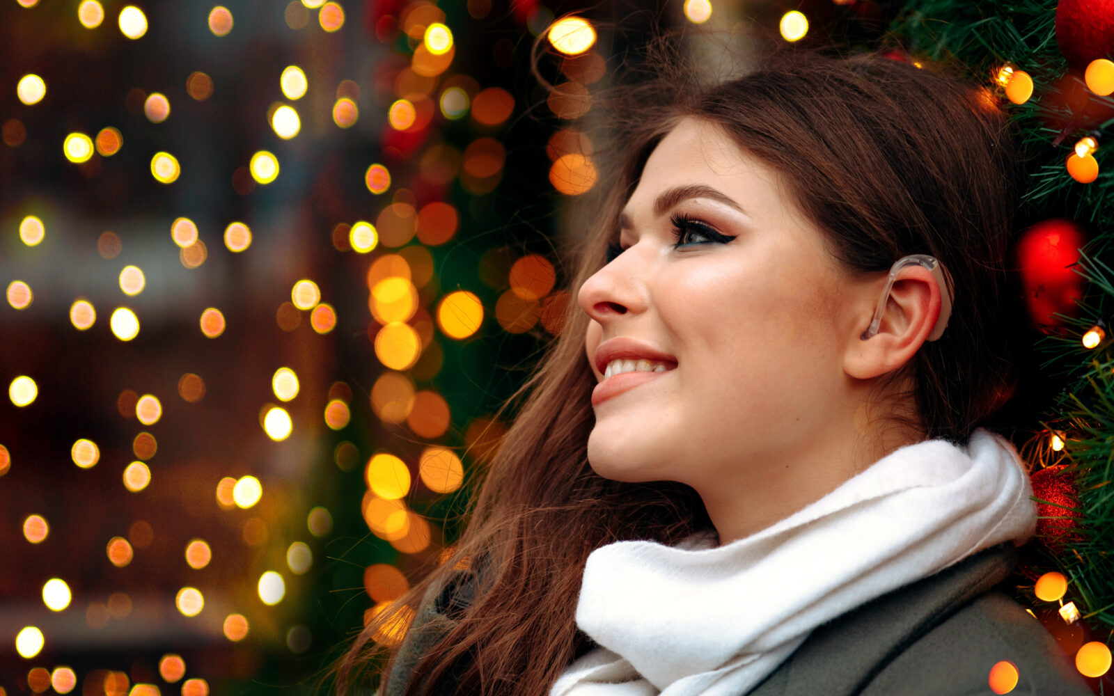 A young woman wearing a behind-the-ear hearing aid smiles while standing outdoors during the holiday season, surrounded by warm, colorful, out-of-focus lights.