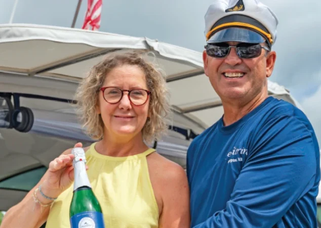 Couple smiling and holding champagne bottle beside their yacht, ready for the christening ceremony