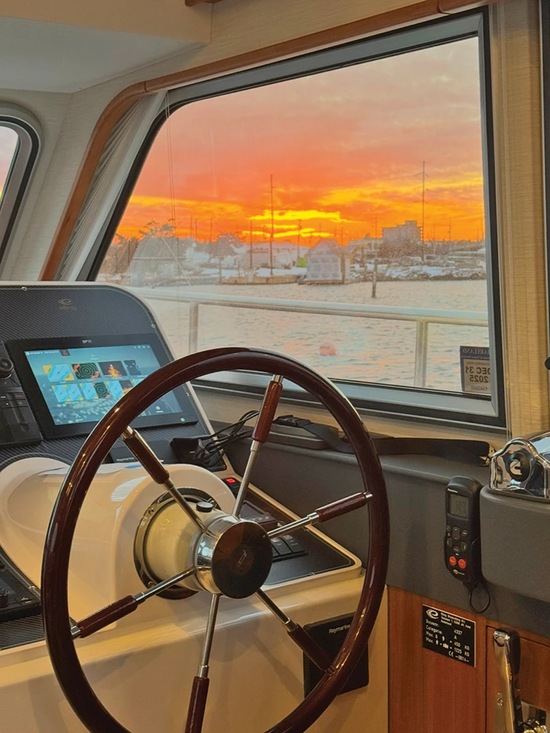 Interior helm station of the Elling E4 with wooden wheel and Raymarine displays, framed by a vivid winter sunset outside
