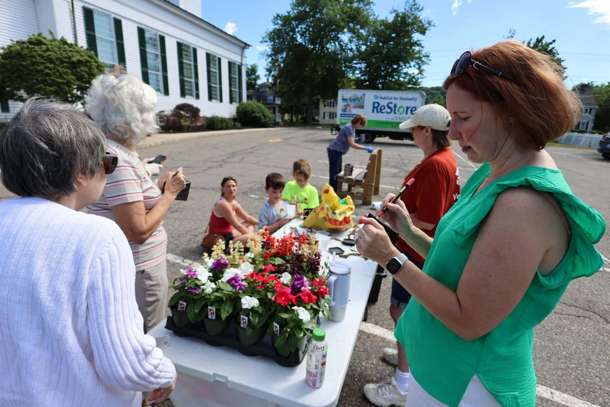Volunteers of all ages potting flowers at a table beside the church, with the Habitat for Humanity ReStore truck behind them