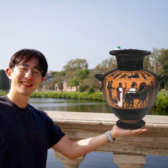 A smiling young man wearing glasses and a dark T-shirt stands on a bridge. He holds his arm out as if to support a Greek vase. The vase is decorated with horses and men in armor carrying shields.
