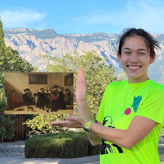 A smiling young woman, standing in a mountainous landscape, gestures toward a painting that appears to float. It represents a 17th-century interior with people, dogs, and paintings. She wears a bright green graphic T-shirt.