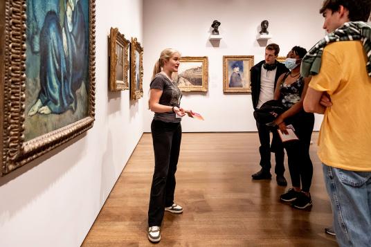 A young woman in a gallery full of elaborately framed paintings speaks in front of a small audience.