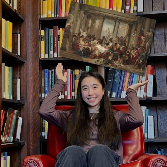 A smiling young woman, wearing a brown long-sleeve shirt and dark pants, sits in a red armchair in a library, gesturing toward a painting that appears to float overhead. In the painting, people in Roman clothes gather in a lofty and statue-filled classical setting.