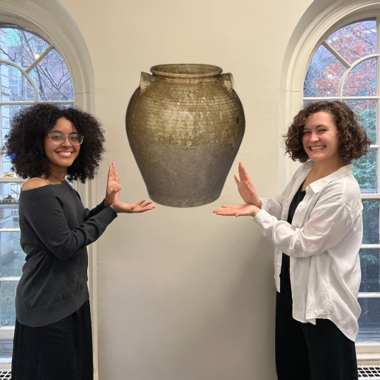 In this photomontage, two smiling young women stand indoors between tall windows, gesturing toward a mottled brown storage jar that appears to float between them.