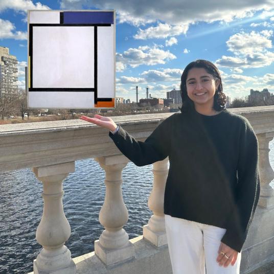 A smiling young woman, wearing a black sweater and white pants, stands on a bridge with a view of a city behind her. She is gesturing toward an abstract painting that appears to float. The painting consists of several flat planes with thick black lines that border mostly white space; three small spaces are filled in with blue, red, and yellow.