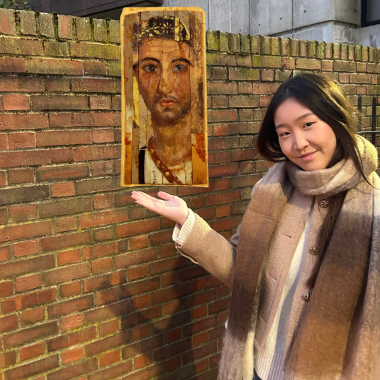 In this photomontage, a young woman, wearing a brown scarf and a beige winter jacket,  gestures toward a portrait that appears to float beside her. The portrait shows a lightly bearded young man and is painted on several slightly mismatched strips of material. 