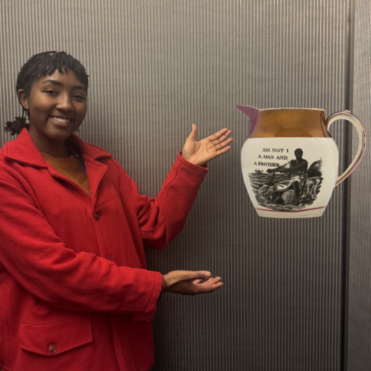 In this photomontage, a smiling young woman gestures toward a jug that appears to float. Printed in black on the jug’s surface is a Black man gesturing toward his chained wrist and the words “AM NOT I A MAN AND A BROTHER.”   