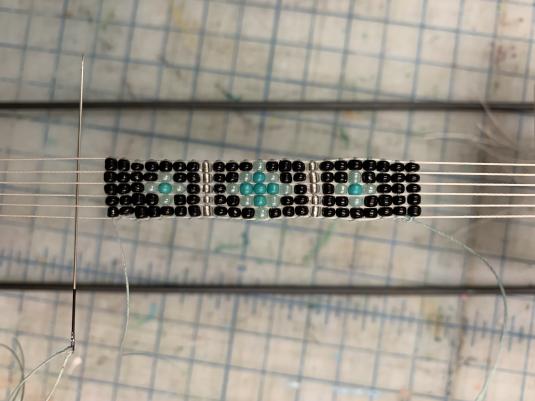 A close-up image of an unfinished bracelet featuring black, white, teal, and silver beads arranged in diamond and floral patterns, positioned on five rows of thick thread.