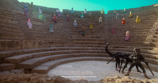 Figures in colorful clothes and masks sit in an amphitheater watching a scorpion-like creature.