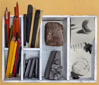 A white wooden box with six cubbyholes contains graphite and charcoal drawing implements, chunks of wood and graphite, and sample drawings.