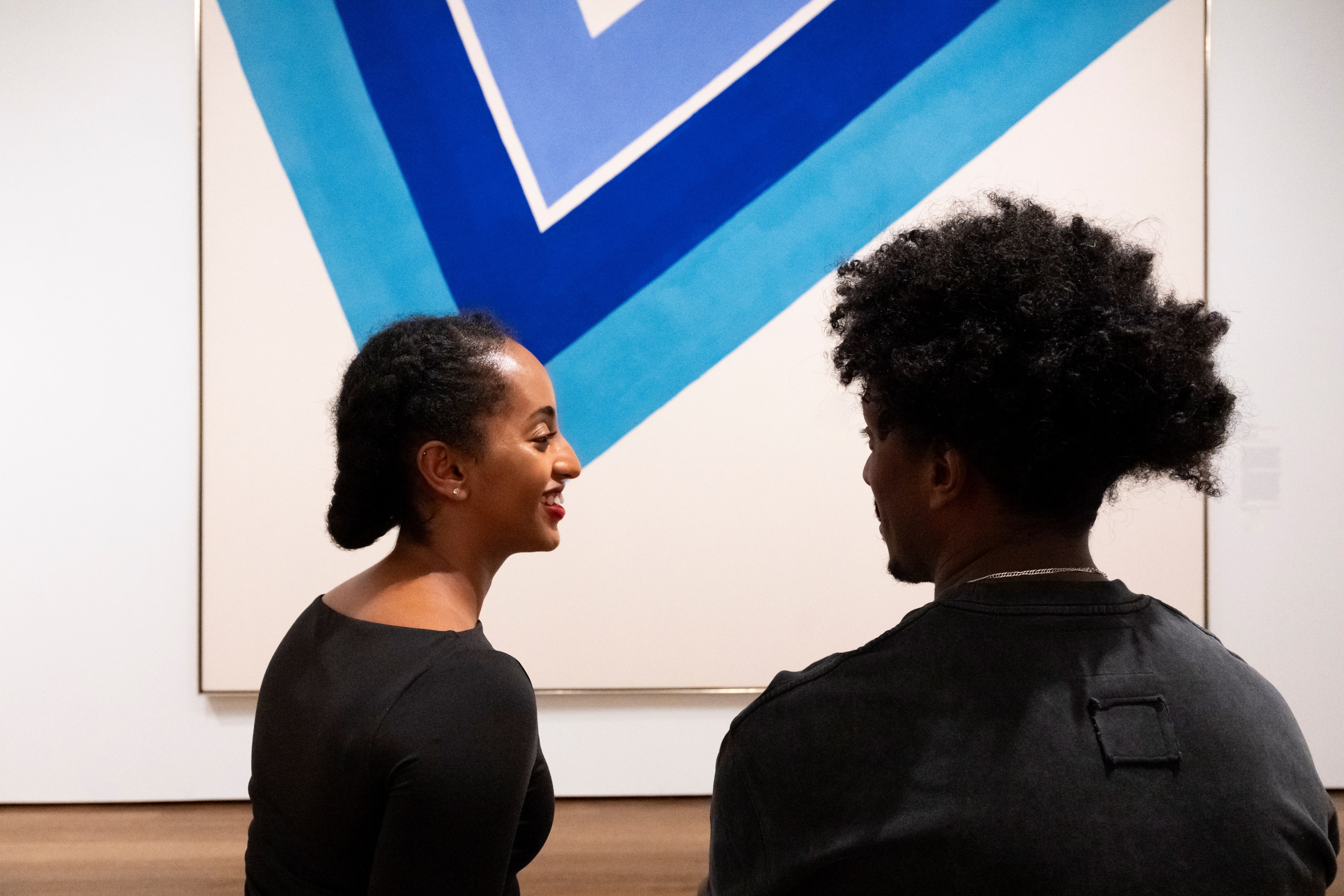 Two museum visitors face one another in front of a large contemporary painting depicting bright blue chevrons.