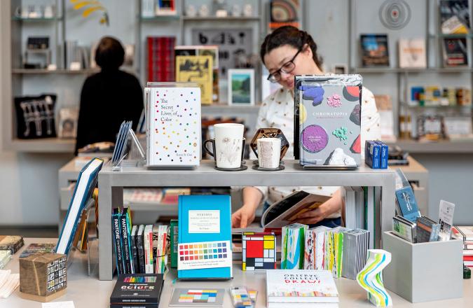 A table arranged with items from the museum shop including books, mugs, and art supplies. Visitors browse the shop in the background.