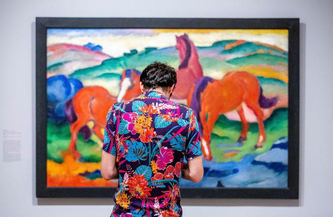 A visitor with a bright, colorful flower print shirt stands in front of a bright, colorful painting of horses.