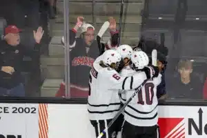 Omaha Mavericks forward Tanner Ludtke is hugged by teammates Zach Urdahl (6) and  Jacob Guevin (10) after scoring the first goal of the night in the first period against the Niagara Purple Eagles during a college hockey match on Friday, October 13, 2023, in Omaha, Nebraska. Photo by John S. Peterson.