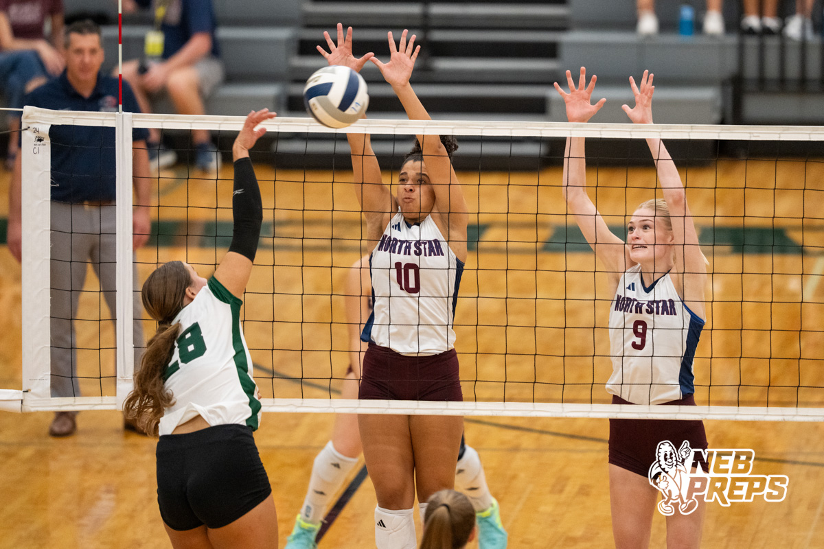 Female volleyball players block at the net during a competitive high school match played indoors on a wooden court.