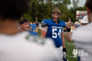 A player in a blue number 54 jersey with a headband and eye black stands on the sideline while another player gestures with a raised finger nearby.