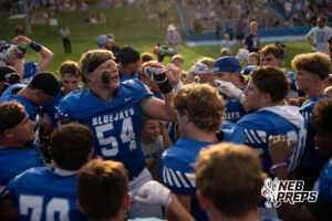 A group of football players in blue jerseys huddle closely together, with one player raising his fist and shouting while surrounded by teammates.