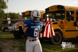 A football player in a blue jersey and helmet carries a flag pole with an American flag draped over it, with school buses parked in the background.