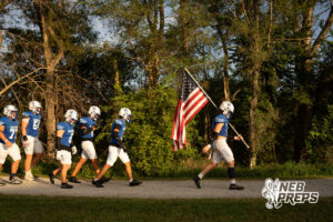 A line of football players in blue jerseys and white pants walks along a gravel path beside trees, led by a player carrying an American flag.