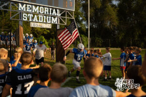 A football player in a blue jersey carrying an American flag leads teammates through a gate under a 'Memorial Stadium' sign, running past a line of young fans reaching out to high-five them.