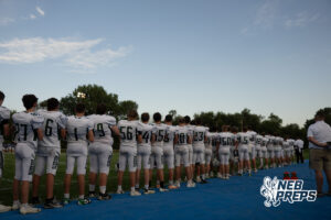 A long line of football players in white uniforms stand shoulder to shoulder along a blue sideline mat, facing away from the camera toward the field.