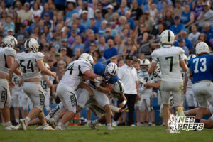 A ball carrier in a blue jersey is being tackled by several defenders in white jerseys near the sideline as a crowd watches from the stands.