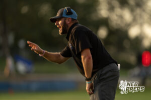 A man in a black polo shirt and a black and blue cap stands bent forward, mouth open, gesturing with one hand while coaching from the sideline.