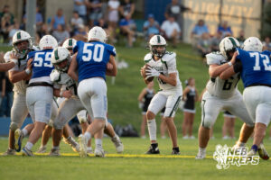 A quarterback in white holds the ball with both hands preparing to pass while linemen in white and blue jerseys engage each other in front of him.