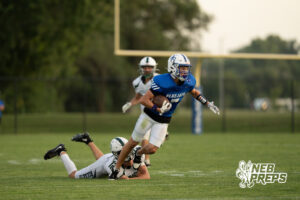 A player in a blue jersey runs with the football past a defender in white lying on the ground, while another defender in white chases from behind near the goal post.