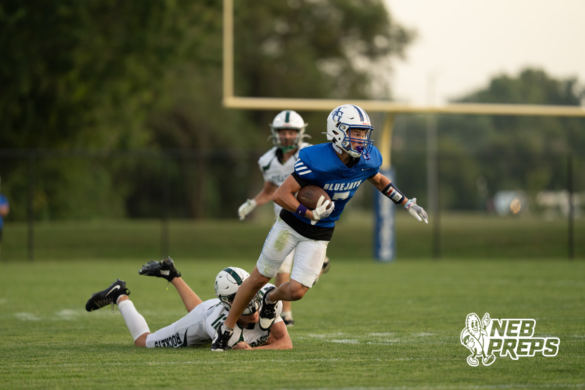 A player in a blue jersey runs with the football past a defender in white lying on the ground, while another defender in white chases from behind near the goal post.