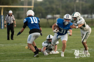 A player in blue carries the football while a defender in white grabs his shoulder pads and another defender falls to the ground nearby.