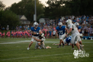 A quarterback in blue is wrapped up by a defender in white while other players converge nearby, with spectators filling the stands behind them.