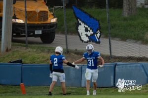 Two players in blue jerseys stand near the end zone padding, one holding a football and reaching toward the other, with a school bus and a fence-mounted mascot logo visible in the background.