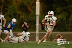 A player in a white jersey runs carrying the football past a fallen teammate and a defender in blue, with a photographer visible in the background.