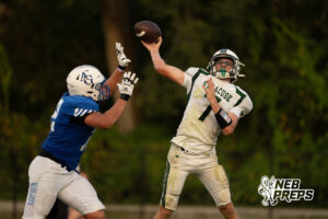 A player in a white jersey throws a football while a defender in a blue jersey reaches up to block the pass.