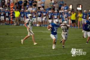 A blue-jerseyed player runs with the football, stiff-arming a white-jerseyed defender, as other players trail behind them on the field near the sideline.
