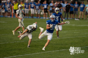A player in blue carries the football past a diving defender in white as another defender and a teammate trail behind, with players lining the sideline in the background.