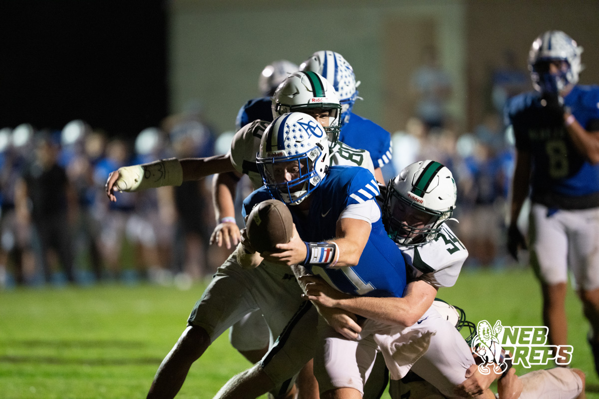 A blue-jerseyed player carrying the football is being tackled by two white-jerseyed defenders as other players watch from the sideline.