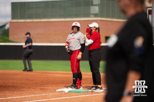 Alexis Jensen stands on first base as a Nebraska staff member holds her batting gear in a college softball game.