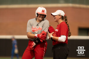A Nebraska player laughs with a Nebraska coach after reaching base in a college softball game.