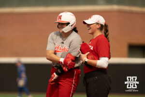 A Nebraska player laughs with a Nebraska coach at first base in a college softball game.