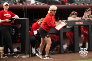 Head coach Rhonda Revelle yells from in front of the Nebraska dugout while holding a clipboard in a college softball game.