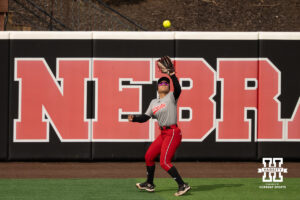 A Nebraska player catches a fly ball in front of the outfield wall in a college softball game.
