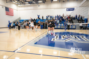 Marian players rush the court to celebrate as fans cheer from the stands in a high school volleyball match.