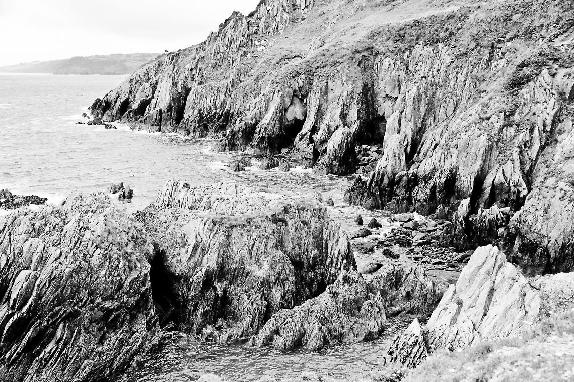Raw coastal enery I forced black and white with a lot of light on this photo to contrast the raw formation of rocks without disturbance of colour.  Wow, This Devon coastline is beautiful!<br />
Taking this shot I actually (so Canon maintenance told me afterwards) got dust blown on my prisma right through the zoom tubes on my lens. The winds were extremely forceful on the height of rock I put my feet on (yet the sea seems quiet here). I did not even know it was possible. I suddenly noticed two  specs appear on my pentaprism. It spoiled my moment:) Geotagged,United Kingdom,coast,devon,formation,raw,rock,sea,wind