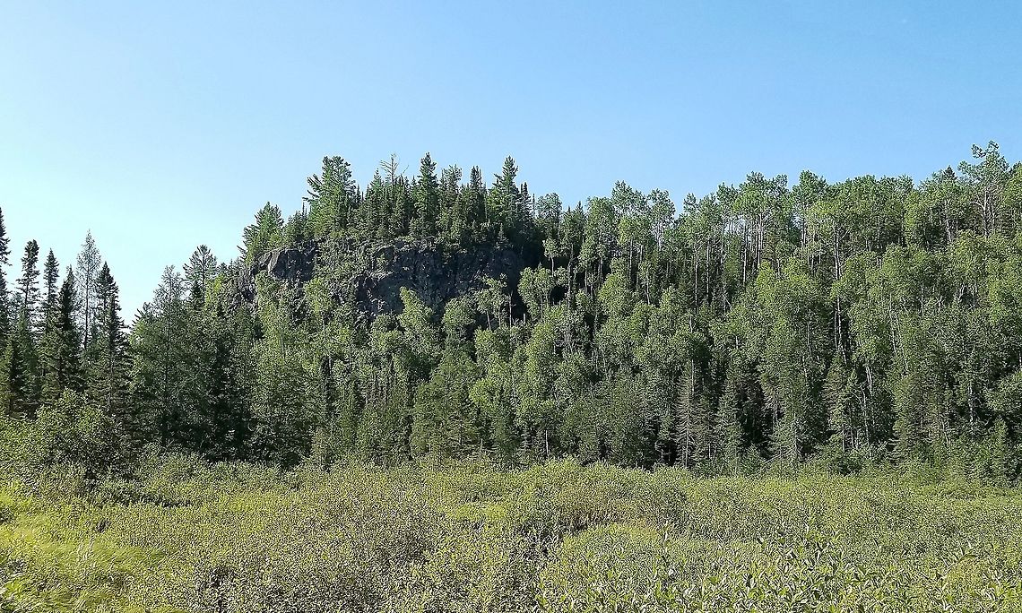 Rock outcrop rising above a shrub carr An outcrop of a dark igneous rock (gabbro? basalt?) forested on its summit with White Pine (Pinus strobus) and Balsam Fir (Abies balsamea). Quaking Aspen (Populus tremuloides) is near the base and on the lower portions of the outcrop. In the foreground is a peatland that is densely covered with Sweet Gale (Myrica gale) a fragrant shrub. A stream (Ninemile Creek) flows throught the peatland. The summit's elevation is about 1,000 feet (~304 meters) above the level of Lake Superior which is 7.3 (~12 km) miles away. Geotagged,Summer,United States,forest,landscape,outcrop,peatland,pine