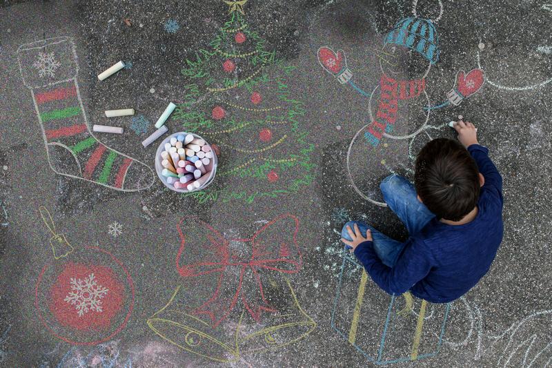 Image: A boy kneels on a sidewalk, drawing with chalk, as various holiday themed chalk drawings surround him.