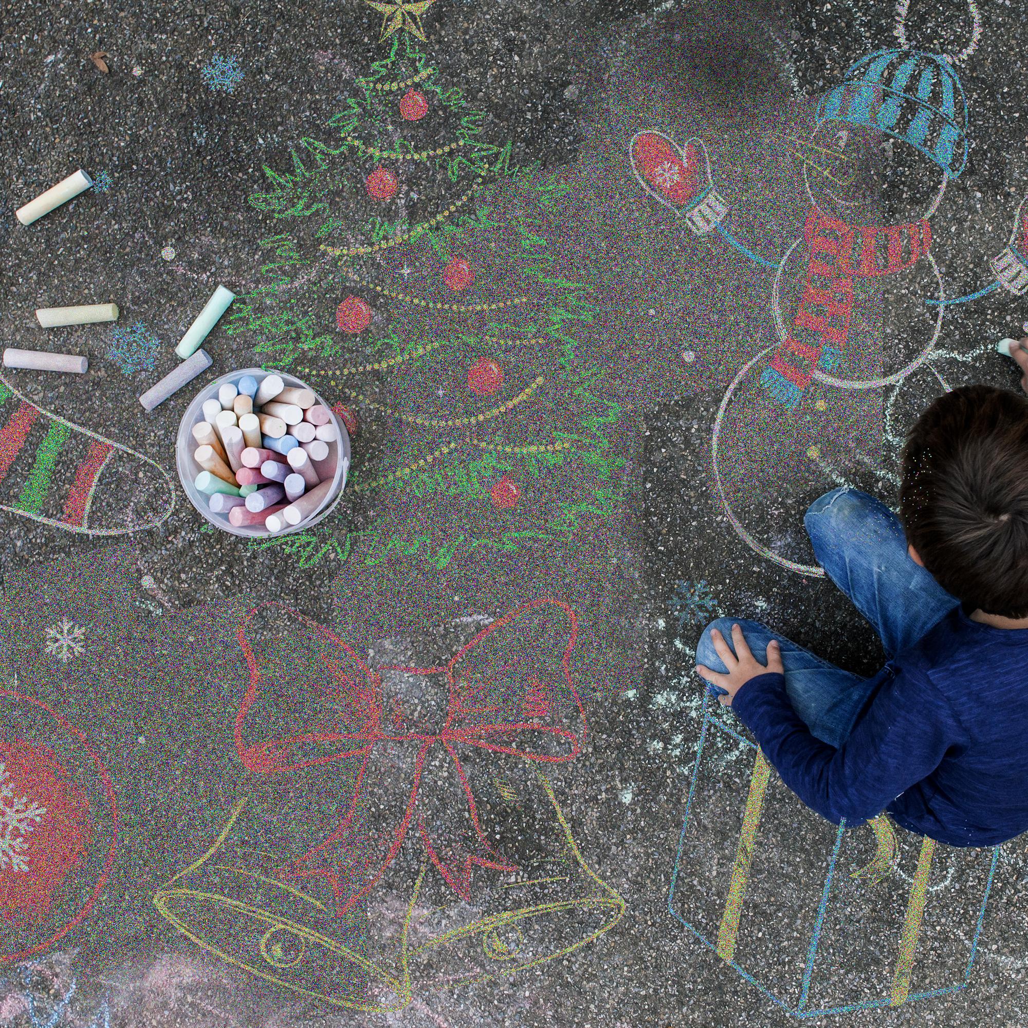 Image: A boy kneels on a sidewalk, drawing with chalk, as various holiday themed chalk drawings surround him.