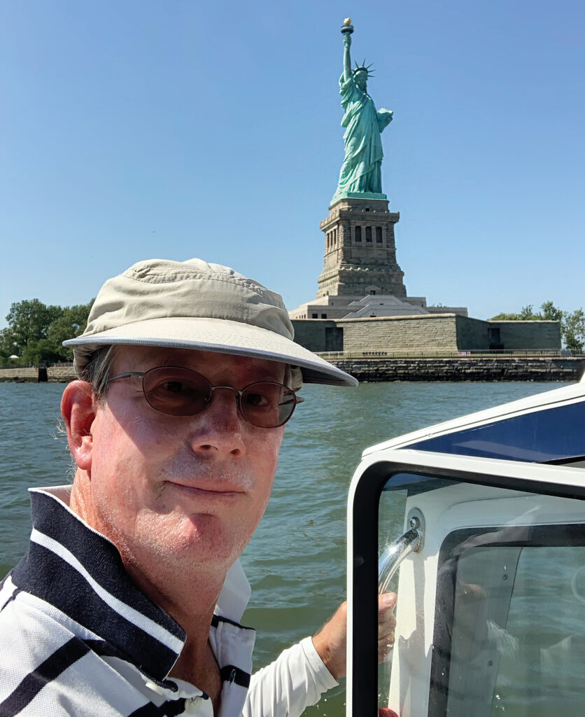 a man in a hat and glasses standing in front of the Statue of Liberty